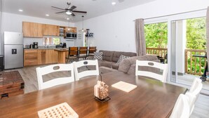 A modern kitchen with wooden cabinets, a dining table with white chairs, and a view of greenery through large windows.