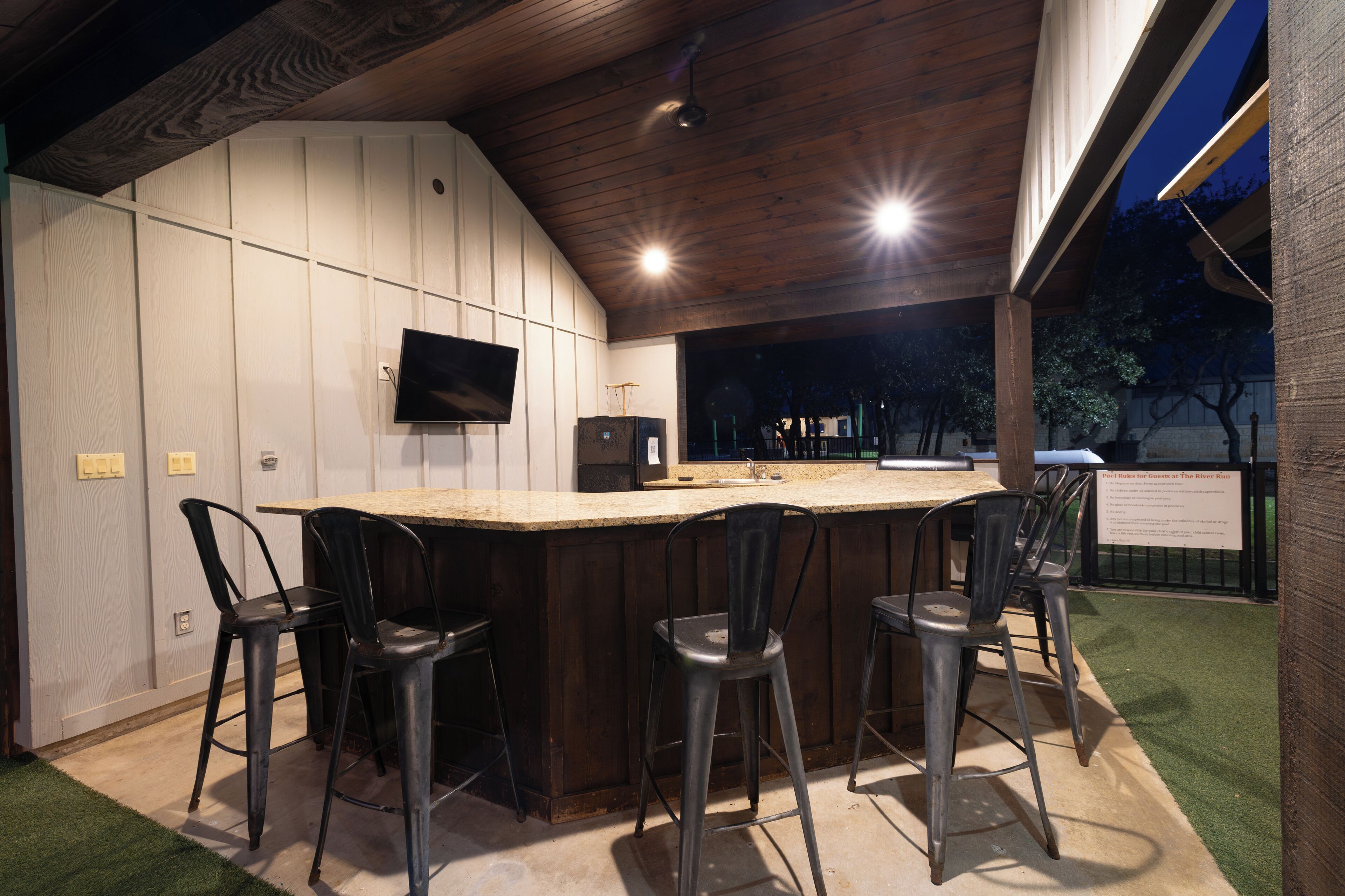 An outdoor bar area with a wooden ceiling, a flat-screen TV mounted on the wall, and four metal bar stools.