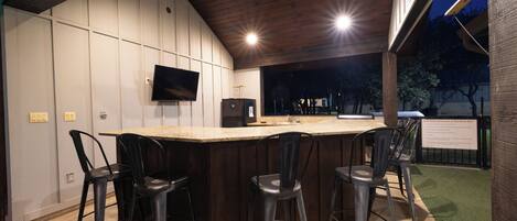 An outdoor bar area with a wooden ceiling, a flat-screen TV mounted on the wall, and four metal bar stools.