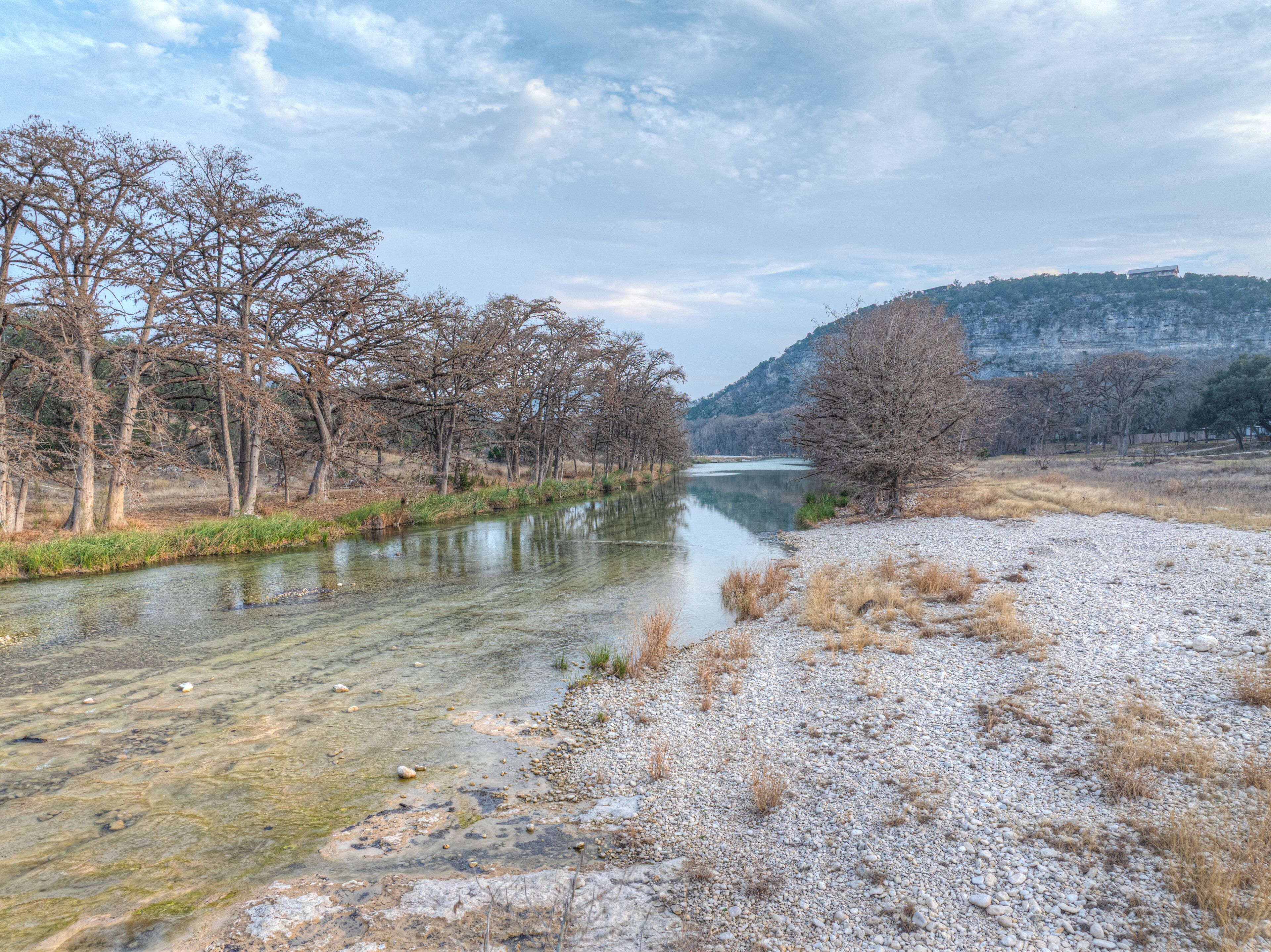 Une rivière bordée d’arbres, un sentier de gravier et une colline en arrière-plan.