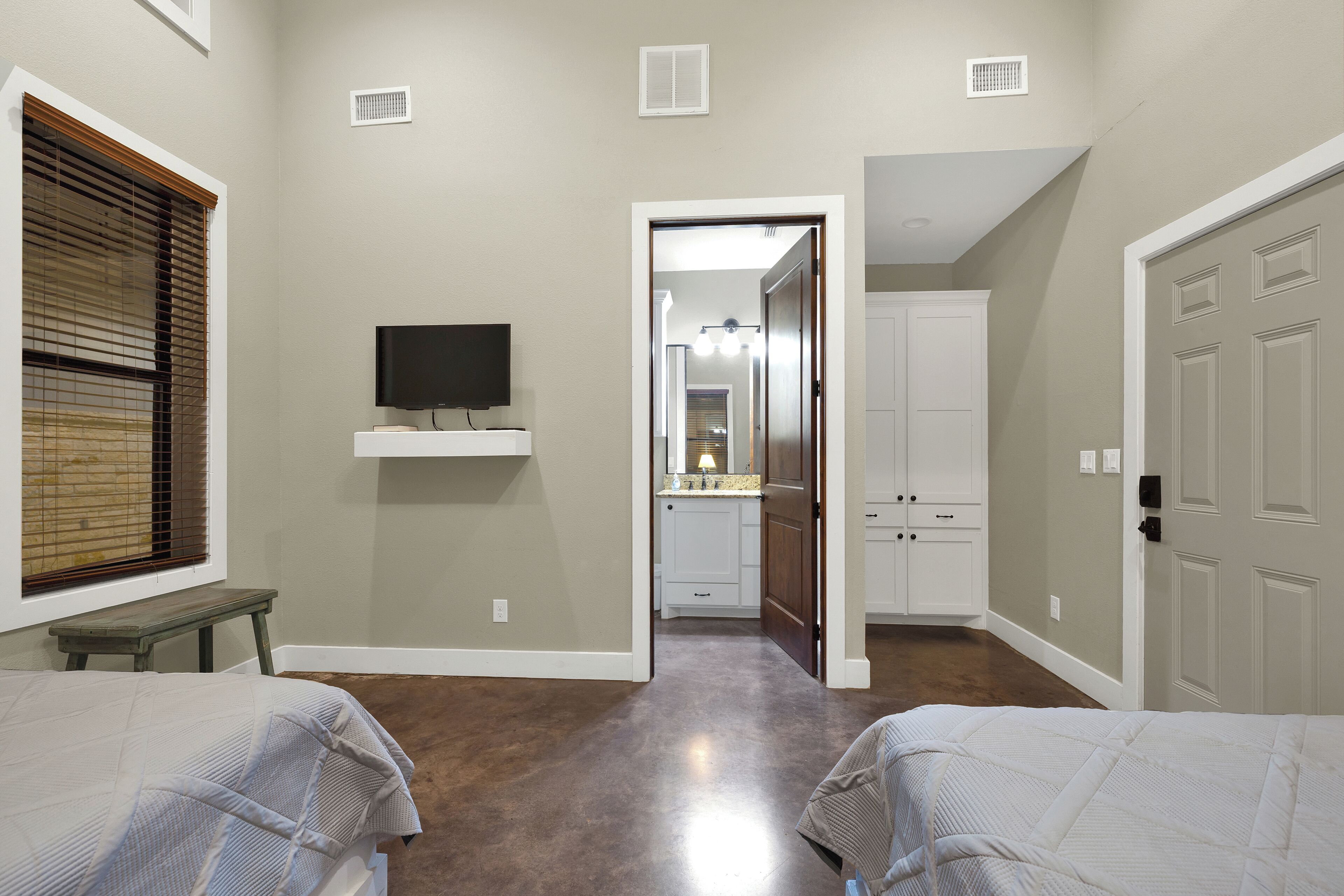 A bedroom with two beds, a TV on a shelf, and a hallway leading to a bathroom.