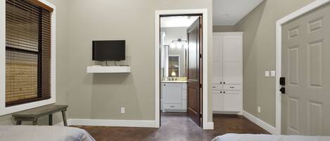 A bedroom with two beds, a TV on a shelf, and a hallway leading to a bathroom.