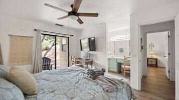 Un dormitorio con cama, escritorio, silla, ventilador de techo y baño con espejo y lavamanos.