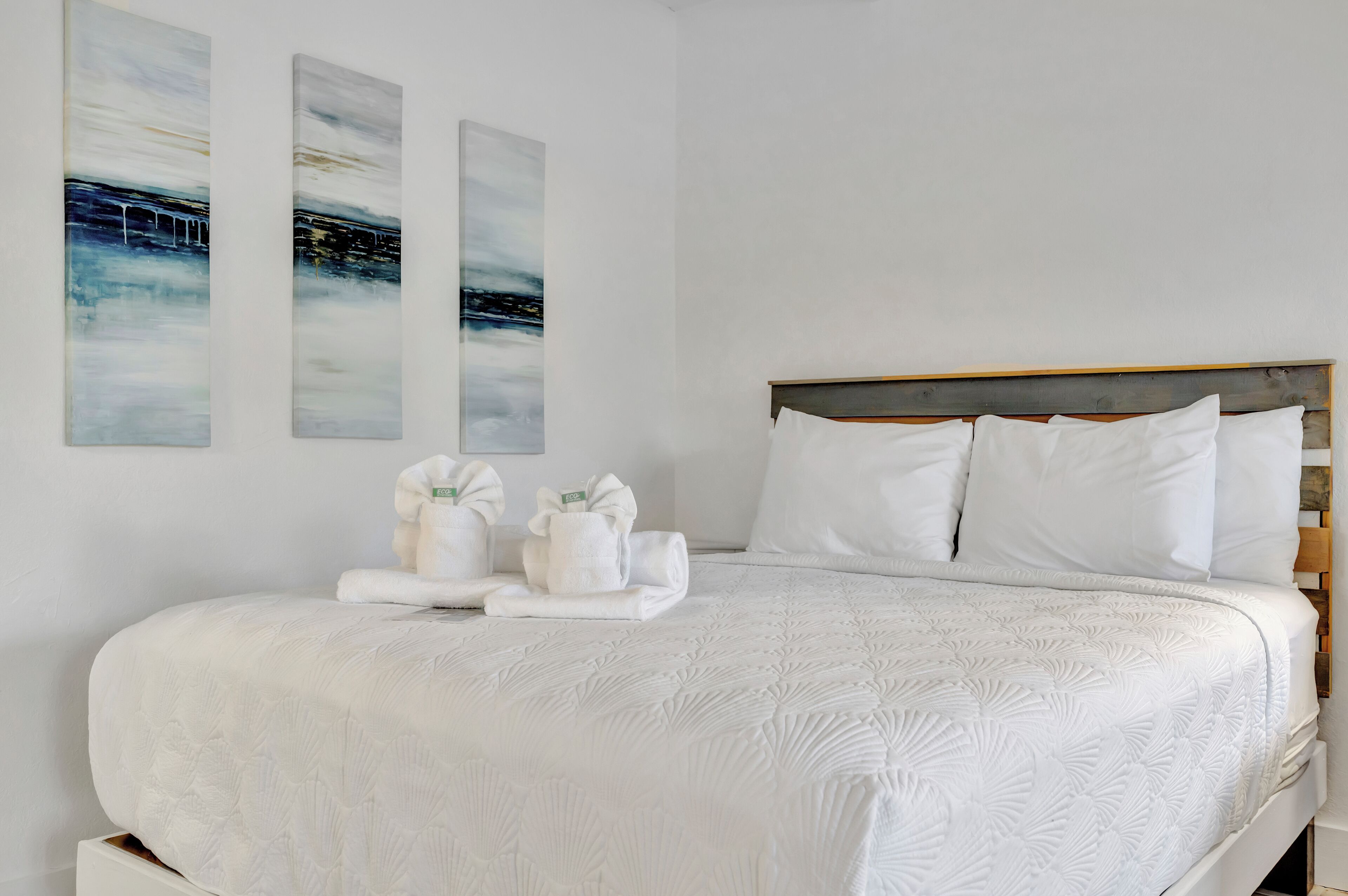Un lit bien fait, avec du linge de lit blanc et des oreillers, une tête de lit en bois et deux tableaux abstraits accrochés au mur.