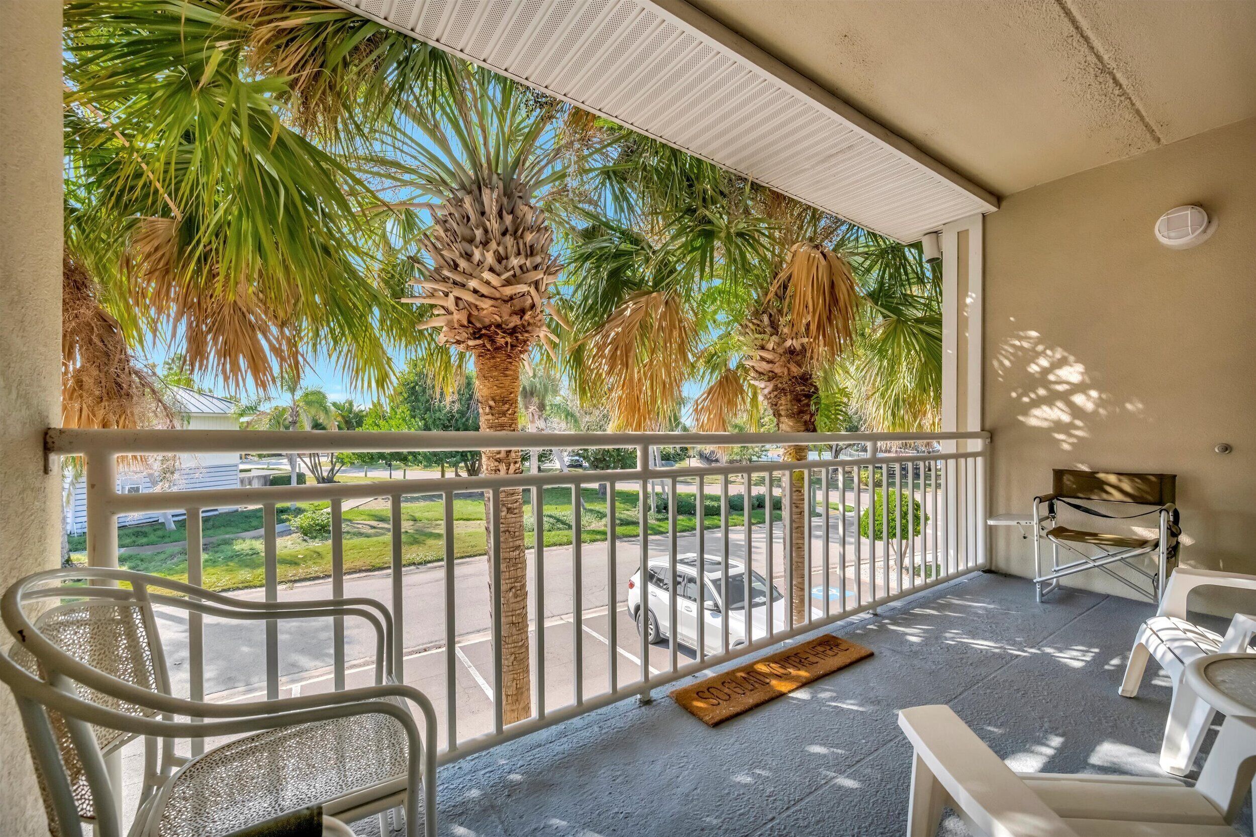 A balcony with a view of palm trees and a parking lot. There are white chairs and a small table on the balcony.