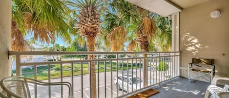 A balcony with a view of palm trees and a parking lot. There are white chairs and a small table on the balcony.