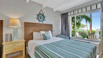 A bedroom with a bed, a bedside table with a lamp, a decorative wall piece, and a window with a view of palm trees.