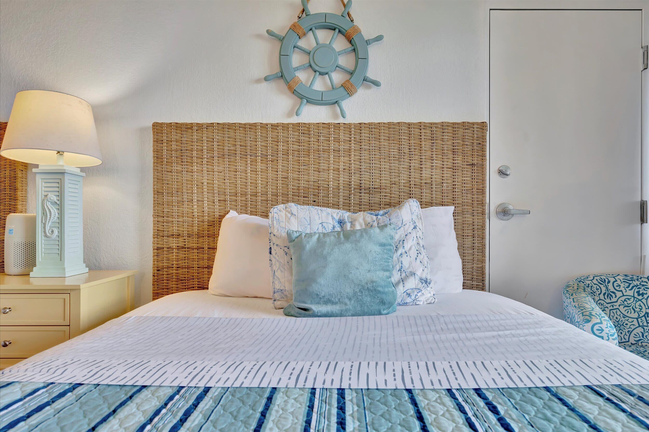 A neatly made bed with a striped bedspread, a headboard with a woven texture, and a wall-mounted ship's wheel decoration.