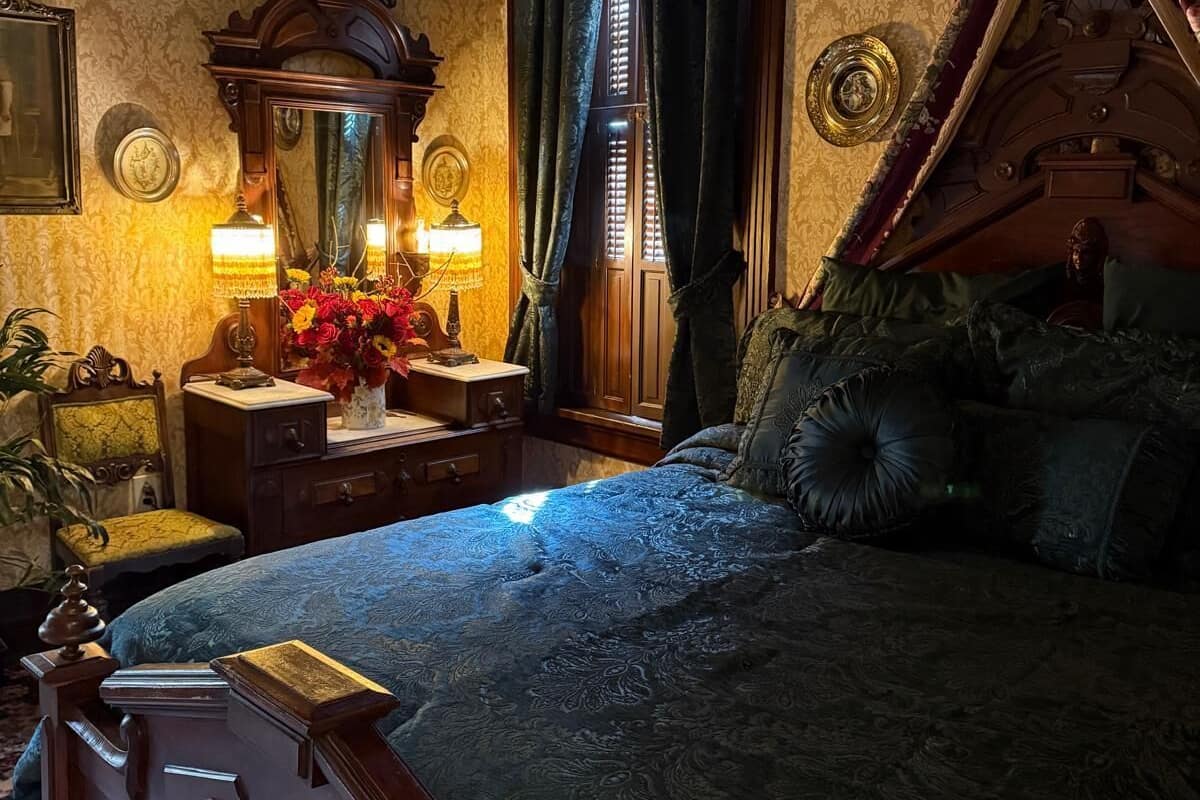 Un dormitorio con cama con dosel, una mesita de noche con lámpara, un espejo y una ventana con cortinas.