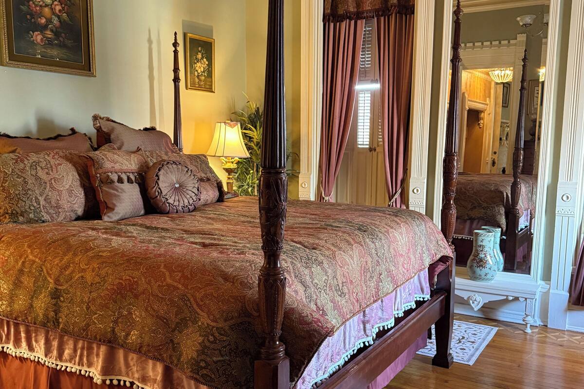 Una cama con dosel, un edredón floral, una mesita de noche con lámpara y un espejo sobre la cama.