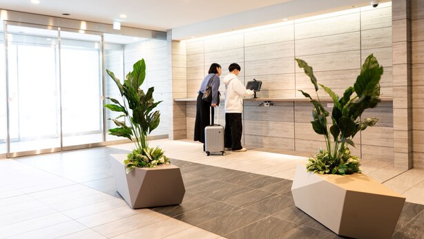 A modern reception area with a check-in counter, two people interacting, and potted plants. - Regale Namba (Osaka)