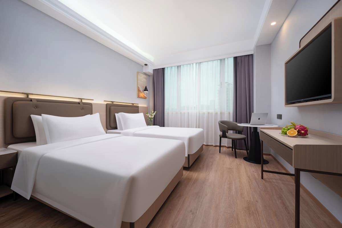 A hotel room with two beds, a desk with a laptop, a flat-screen TV, and a small table with fruit.