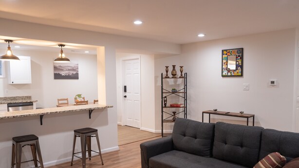 A modern living room with a grey sofa, wooden floor, kitchen area with a counter and stools, and a decorative shelf with vases.