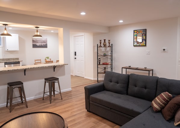 Una sala de estar moderna con un sofá gris, piso de madera, zona de cocina con mostrador y taburetes, y un estante decorativo con jarrones.