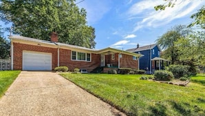 A two-story brick house with a garage, a front yard with grass and shrubs, and a blue house in the background.
