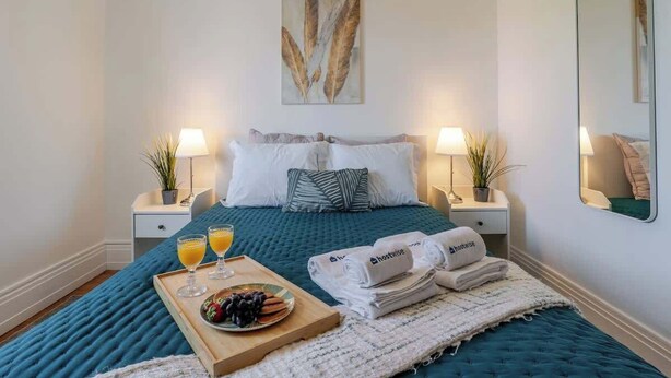 A neatly made bed with a teal duvet, two bedside lamps, a framed artwork above the bed, and a tray with fruit and juice on the side. - Bright & Spacious Flat in Porto by HostWise (Porto)