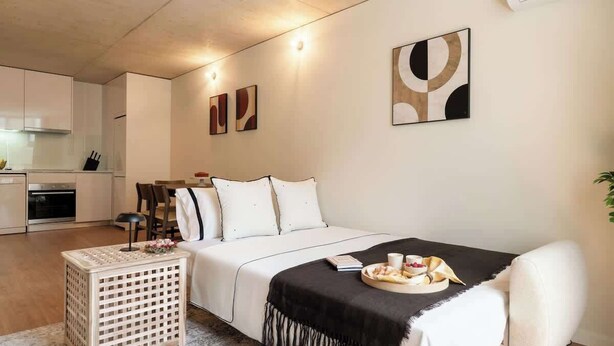 A modern bedroom with a large bed, a bedside table, and a small side table. There is a kitchen area visible in the background. - Modern & Stylish Flat by HostWise (Porto)