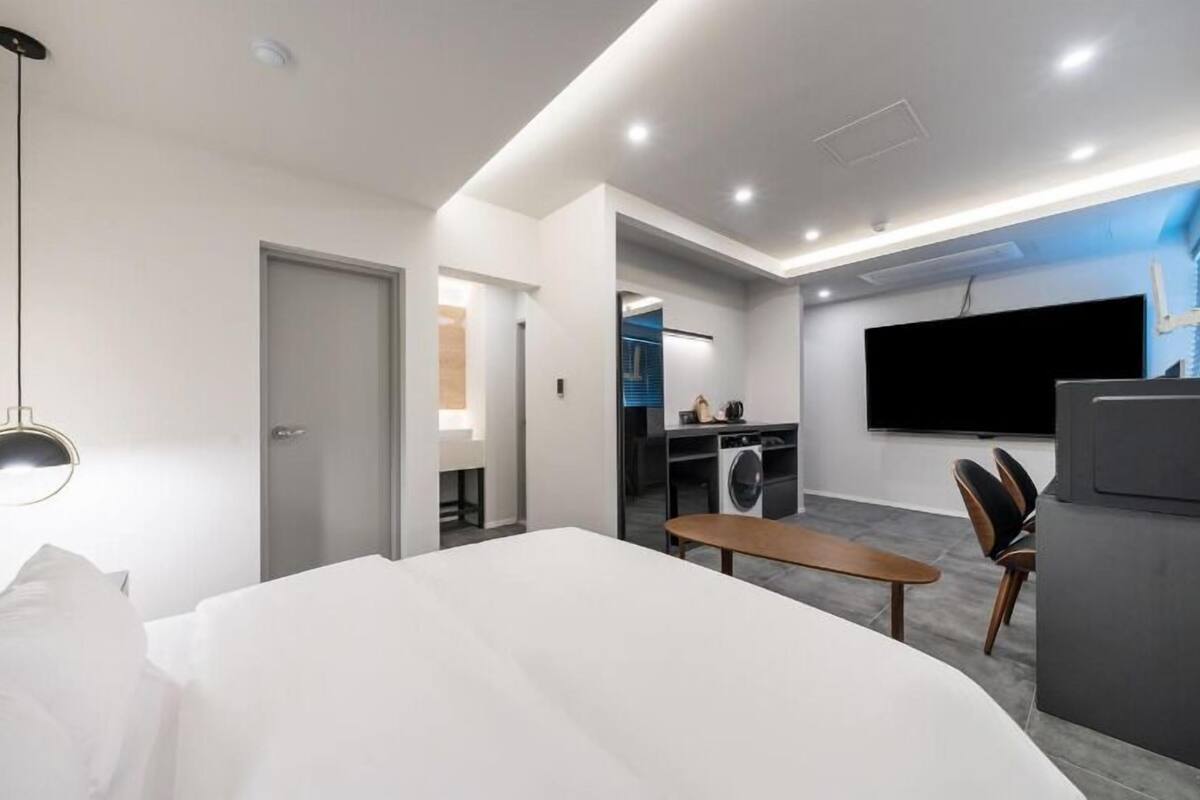 A modern hotel room with a large bed, a dining table, a TV, and a laundry area.