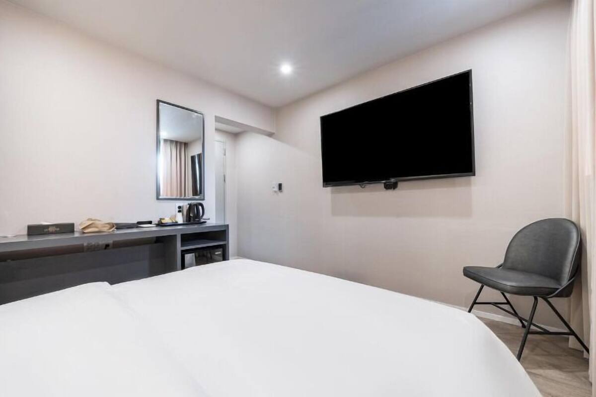 A hotel room with a bed, a desk with a telephone, a chair, a flat-screen TV mounted on the wall, and a mirror.
