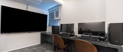 A modern office space with a large flat-screen TV, two computer monitors, a keyboard, and two wooden chairs.