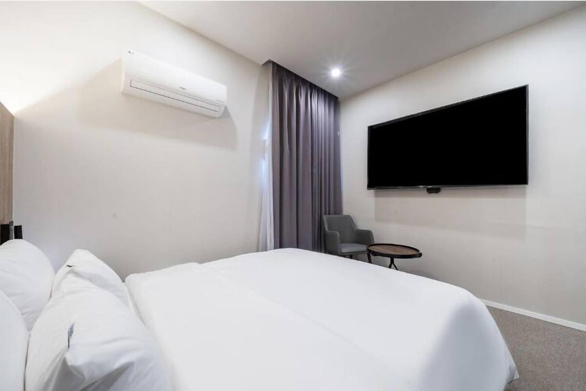 A hotel room with a bed, a chair, a small table, a flat-screen TV mounted on the wall, and an air conditioning unit.