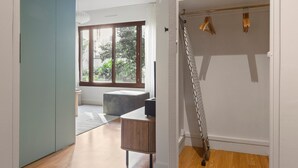 Une chambre moderne avec une grande fenêtre, un plancher en bois, un canapé gris et une armoire blanche où sont suspendus des vêtements.