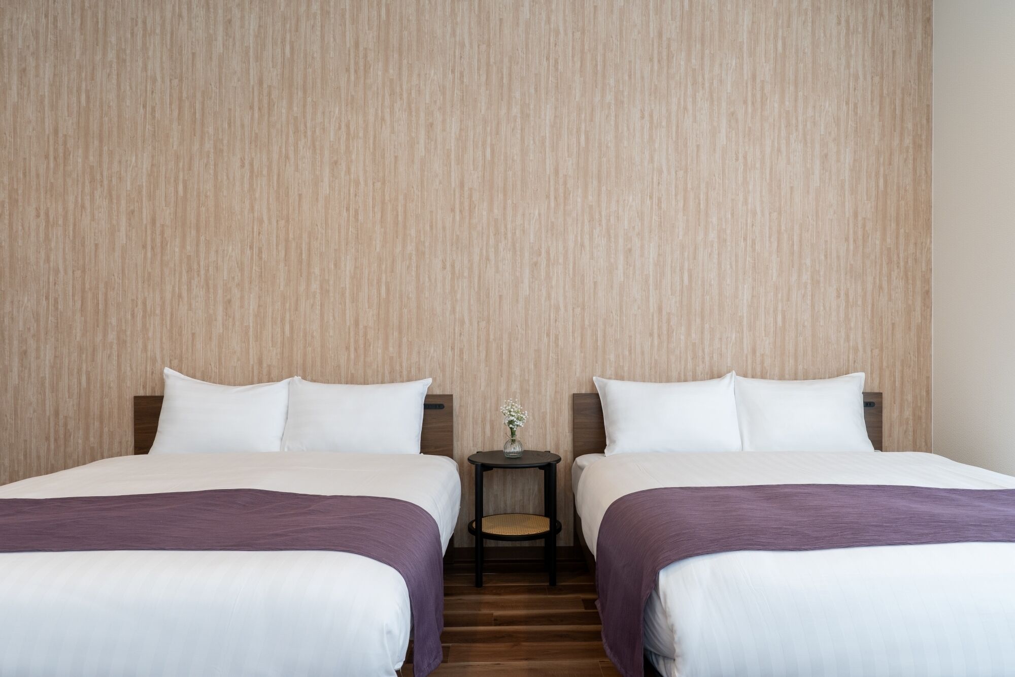 A hotel room with two beds, a small table with a vase of flowers, and a wooden floor.