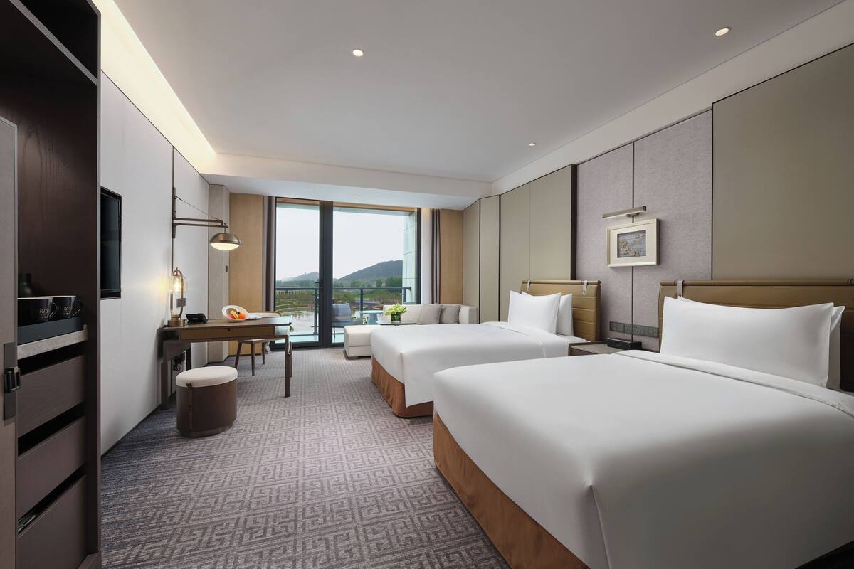 Habitación de hotel con dos camas, un escritorio, una silla, un televisor y un balcón con vistas a las colinas.