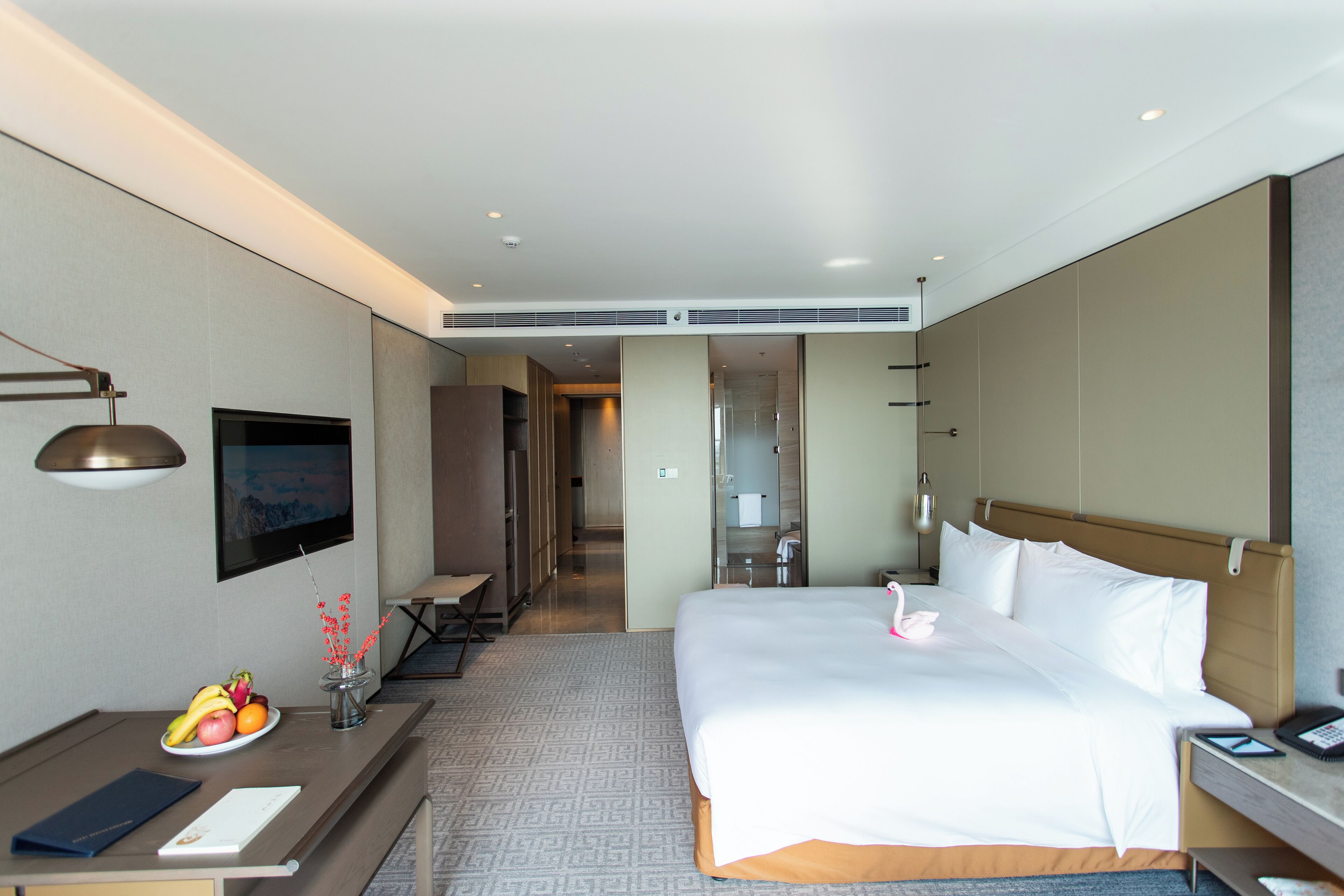Habitación de hotel con una cama grande, un escritorio con frutero y un jarrón con flores, y vista al pasillo.