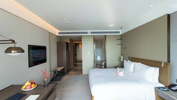 Habitación de hotel con una cama grande, un escritorio con frutero y un jarrón con flores, y vista al pasillo.