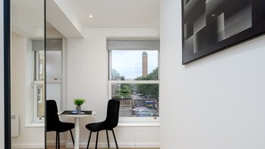A modern room with a large window, a small table, two black chairs, and a potted plant. - GuestReady - Cosy studio in central Greenwich (London)