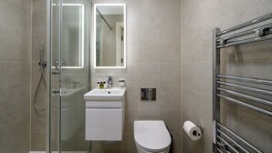 Un baño moderno con cabina de ducha de vidrio, un lavamanos blanco con un espejo encima y un inodoro con dispensador de toallas de papel.