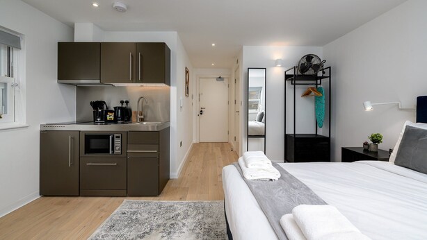 Habitación de hotel compacta con kitchenette, cama, mesita de noche, espejo y armario.