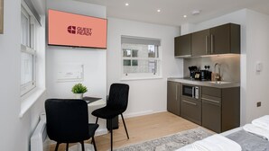 Habitación de hotel compacta con kitchenette, zona de comedor con dos sillas, cama con edredón blanco y vista a la ciudad desde la ventana.