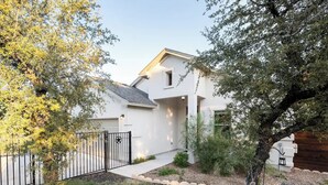 Ein modernes, zweistöckiges Haus mit weißer Fassade, einem schwarzen Metallzaun und einem gepflegten Garten mit verschiedenen Sträuchern und einem Baum.