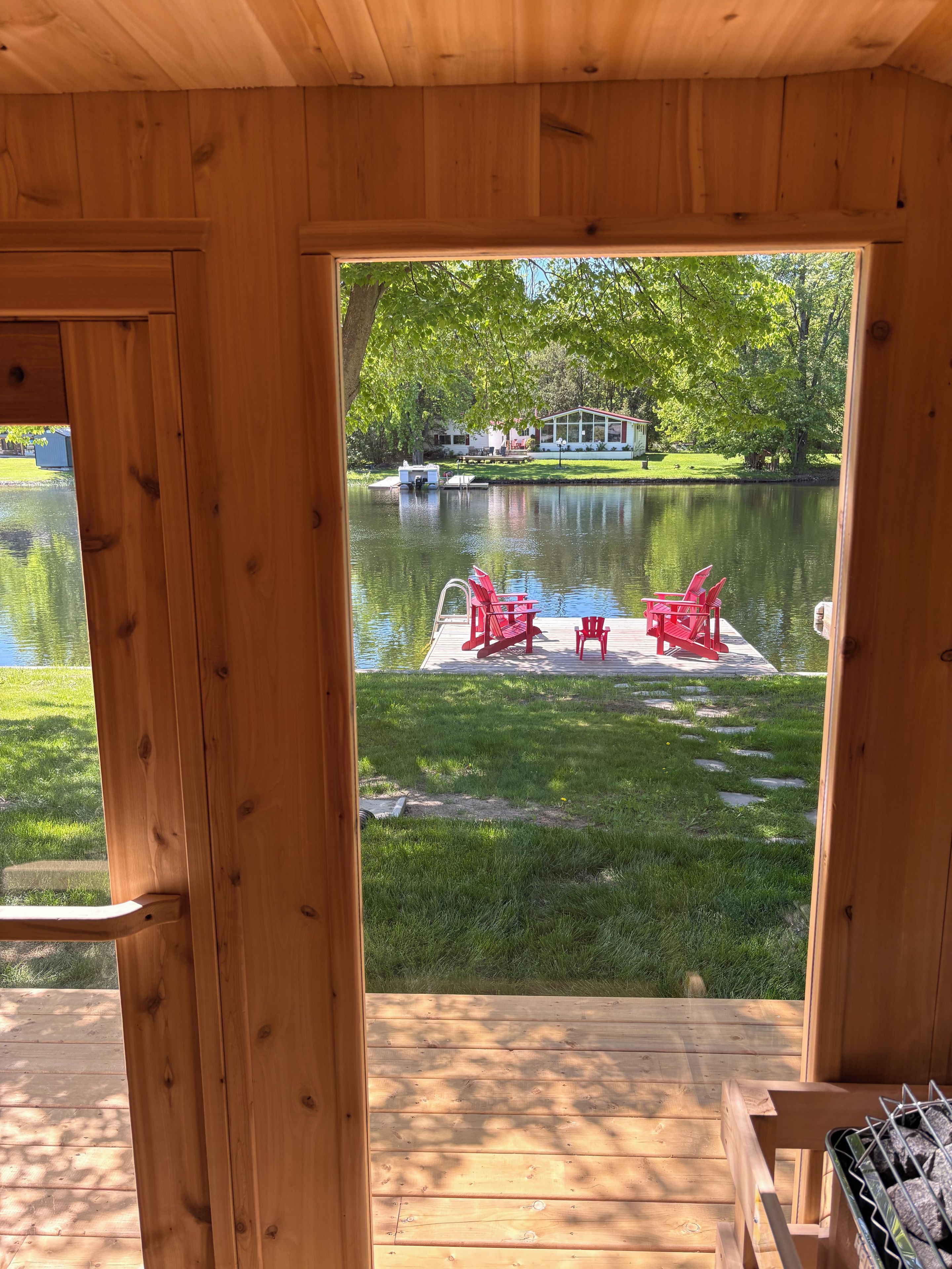 A wooden deck with a view of a lake, red chairs, and a grill.