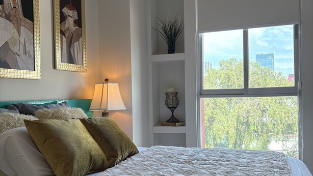 A bedroom with a bed, pillows, a nightstand, a lamp, a window, and wall art. - Cozy apartment close to everything CDMX! (Ciudad de México)