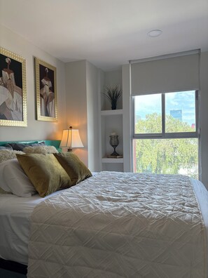 Ein Schlafzimmer mit Bett, Kissen, Nachttisch, Lampe, Fenster und Wandbildern.