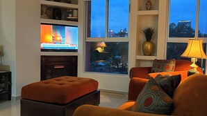 A modern living room with a sofa and ottoman, a TV, and a view of the city at dusk. - Cozy apartment close to everything CDMX! (Ciudad de México)