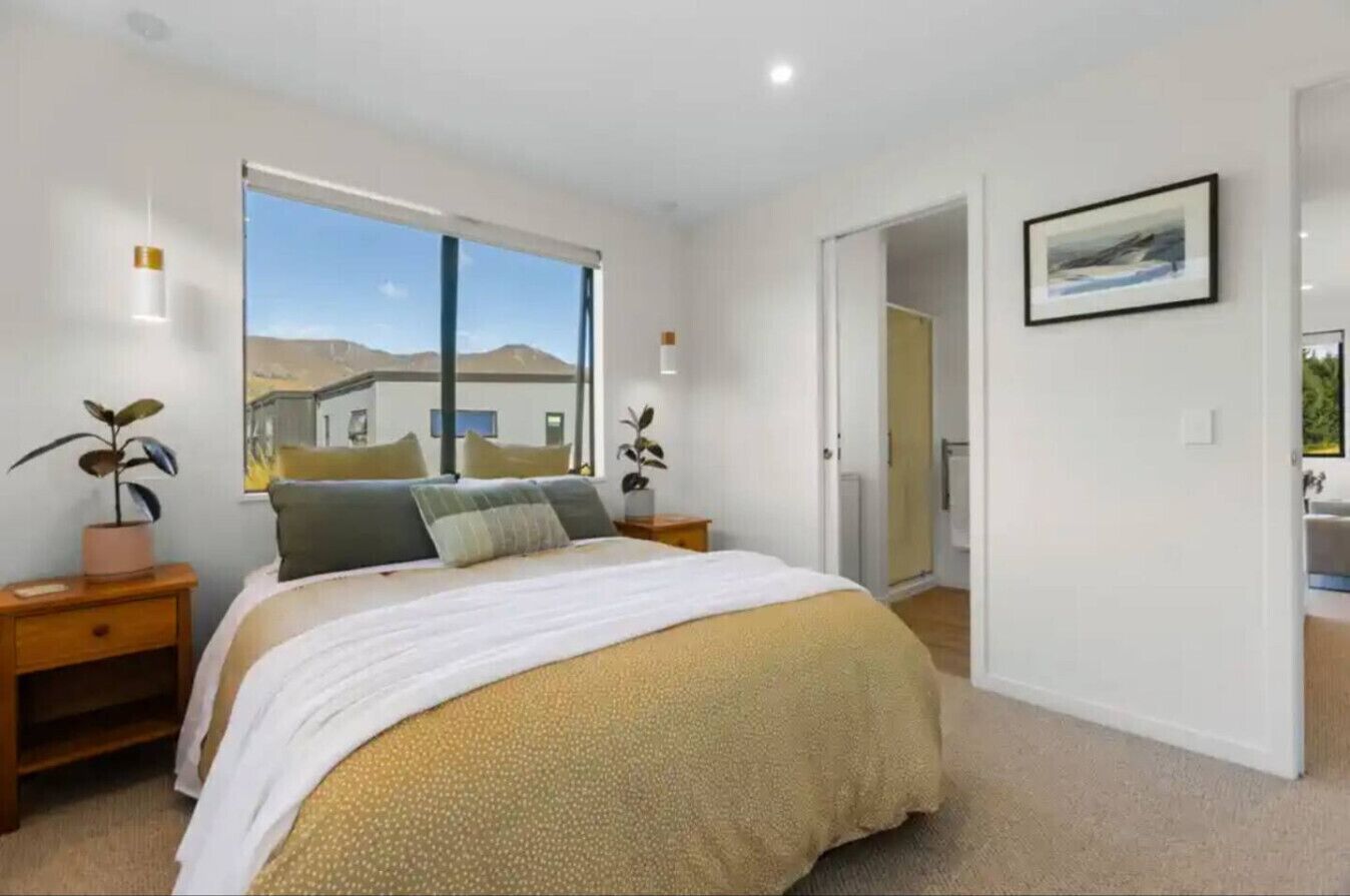 A bedroom with a large bed, a window with a view of a building, a door leading to another room, and a framed picture on the wall.