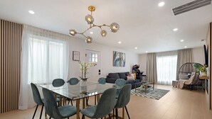 A modern living room with a glass dining table, grey upholstered chairs, a dark sofa, a hanging light fixture, and a gaming console.