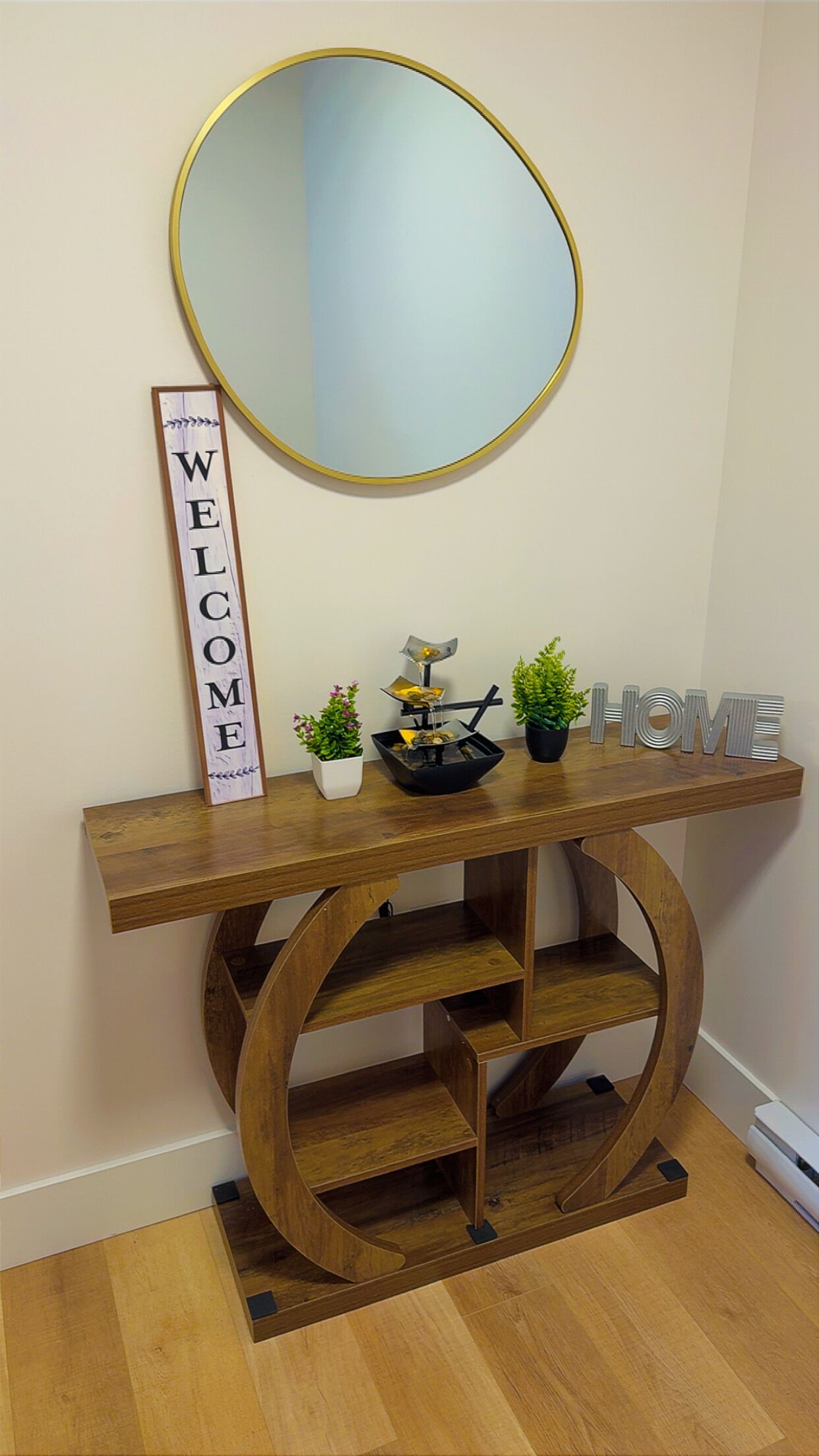Un tavolo consolle in legno con uno specchio sopra, un cartello con la scritta 'WELCOME' e oggetti decorativi tra cui piante e una piccola scultura.
