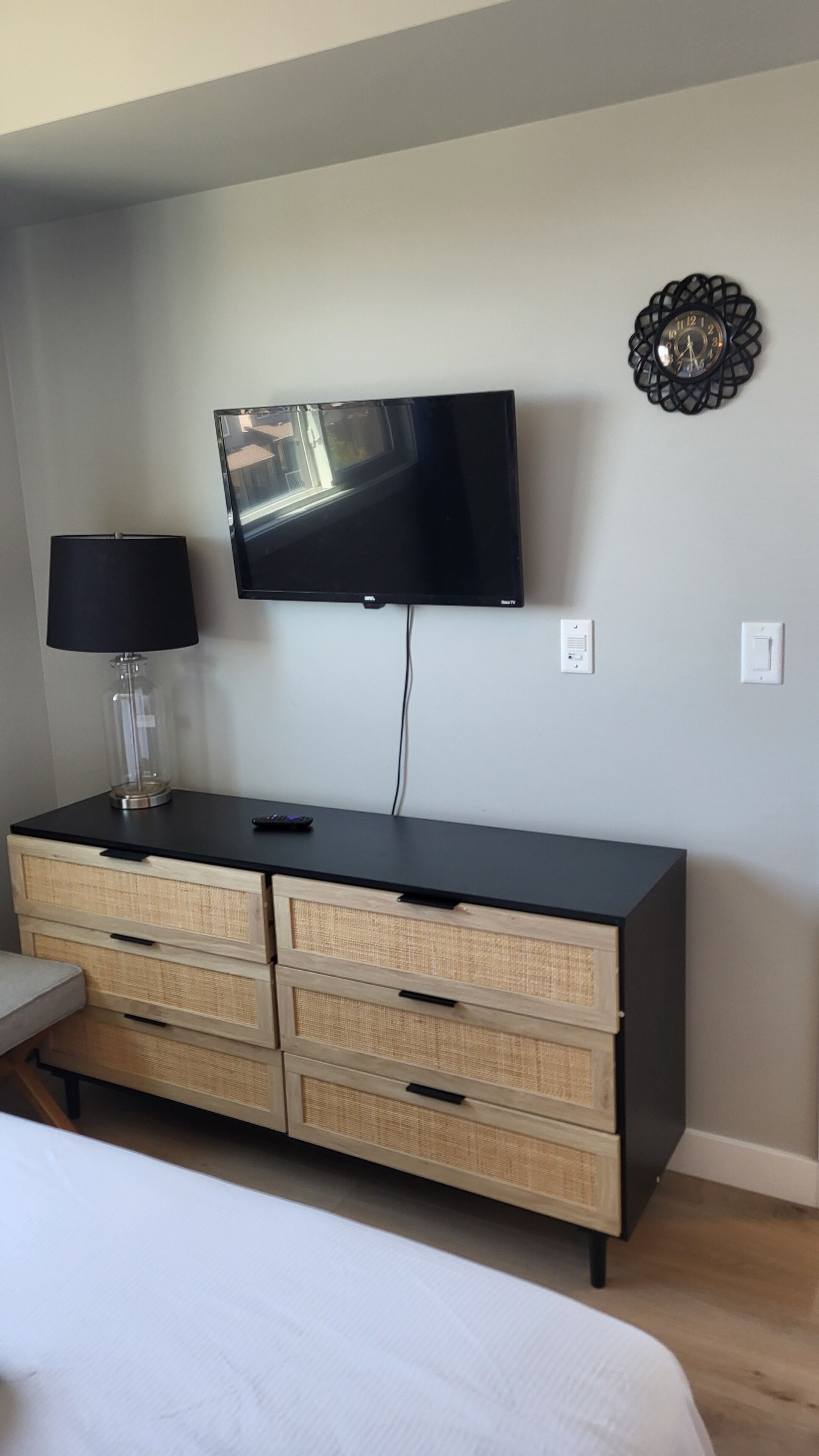 A bedroom with a bed, a TV mounted on the wall, a bedside lamp, and a wooden dresser with drawers.