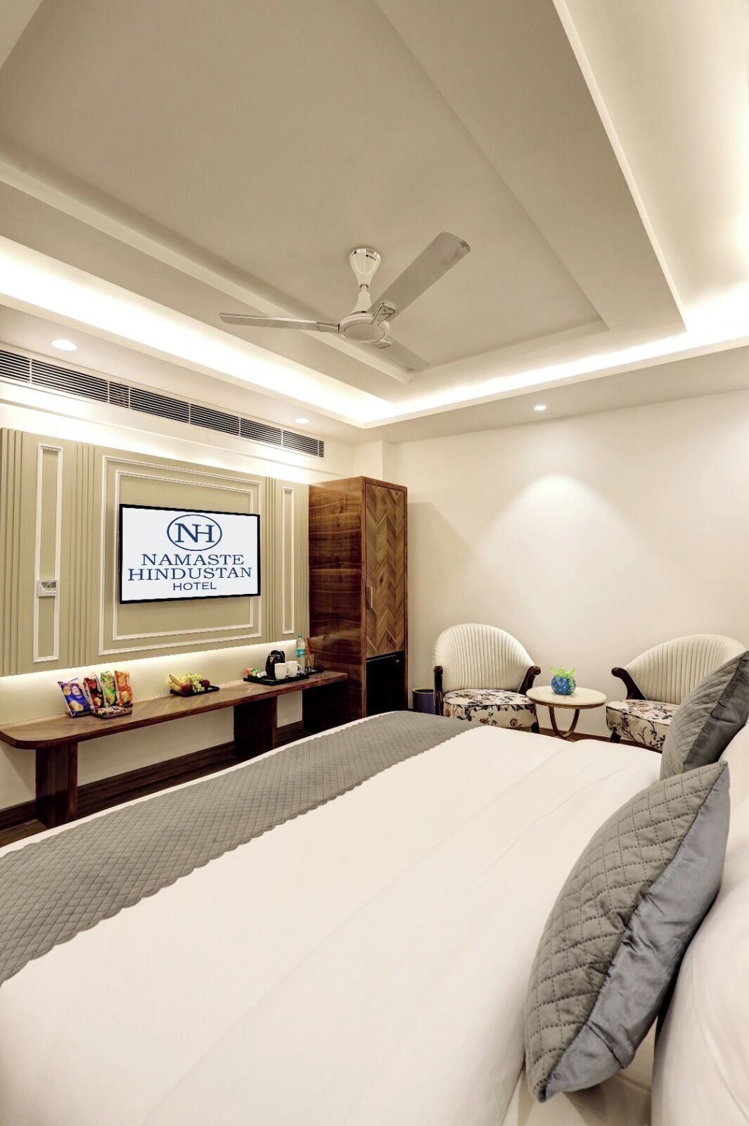 Habitación de hotel con cama, zona de estar con mesa y televisor de pared mostrando el nombre del hotel.