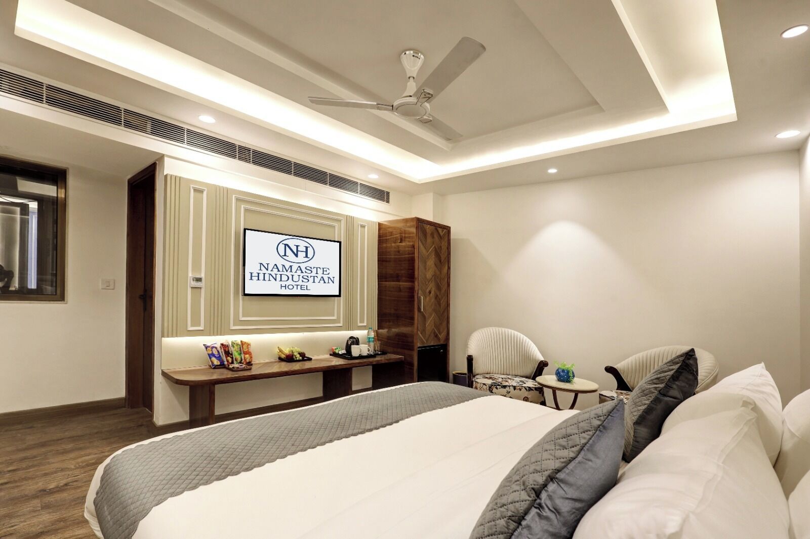 Une chambre d’hôtel équipée d’un lit, d’un bureau, d’une chaise, d’un ventilateur de plafond et d’une enseigne indiquant « Namaste Hindustan Hotel ».