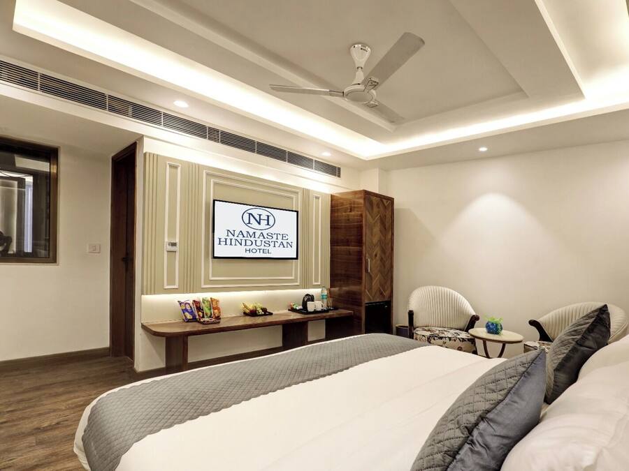 Una habitación de hotel con una cama, un escritorio, una silla, un ventilador de techo y un cartel del Hotel Namaste Hindustan.