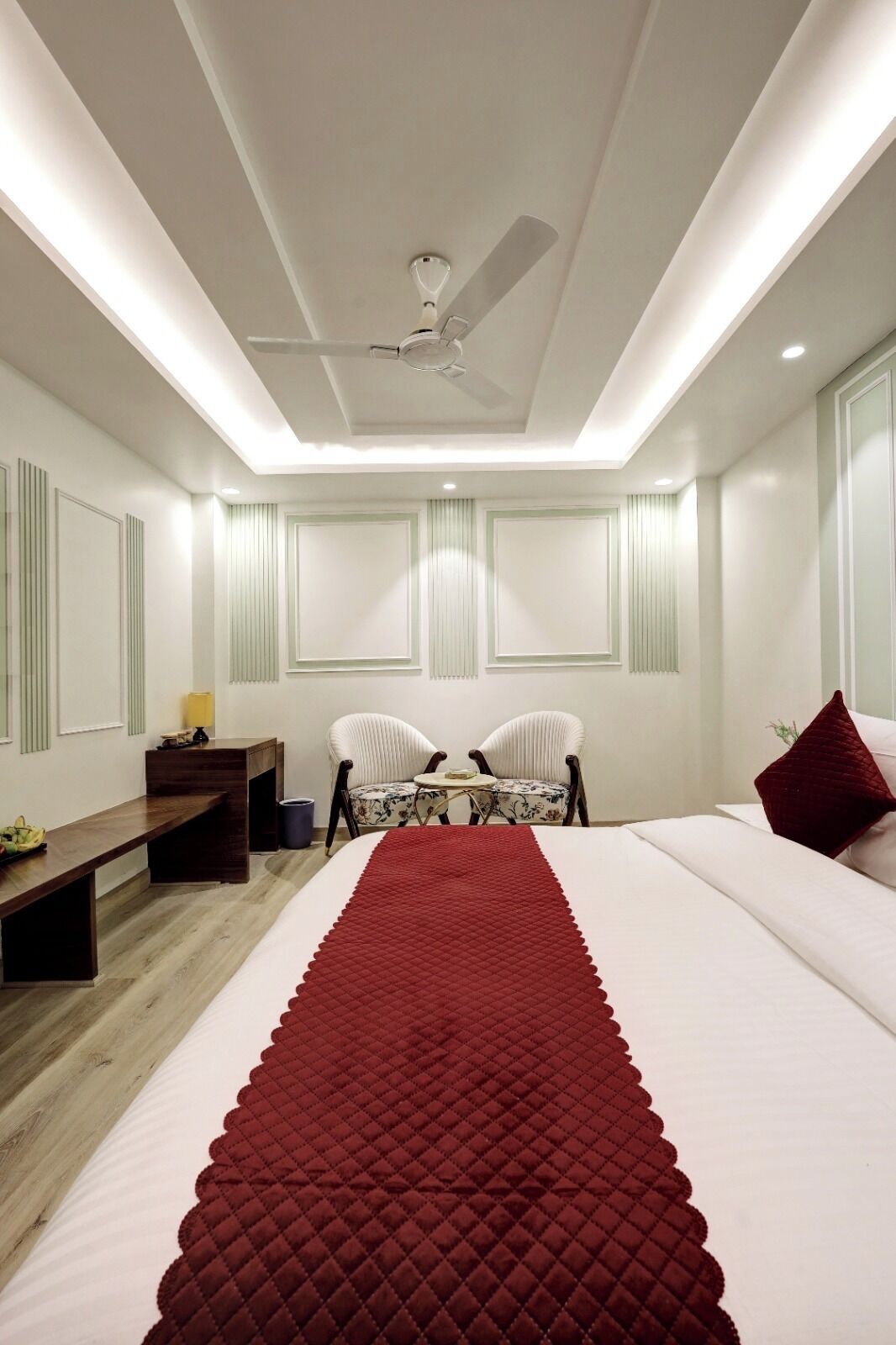 A modern hotel room with a large bed, a desk, two chairs, a ceiling fan, and a red accent wall.