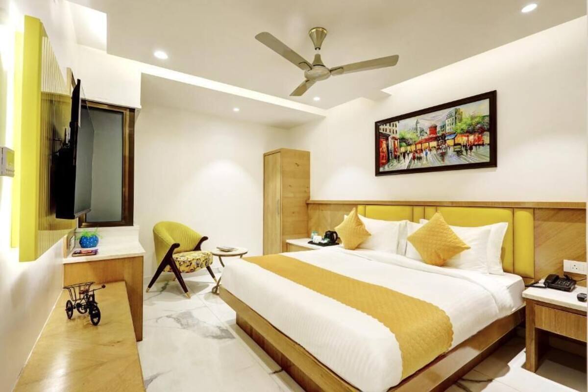 Quarto de hotel com cama, escrivaninha, cadeira, televisão, ventoinha no teto e um quadro na parede.