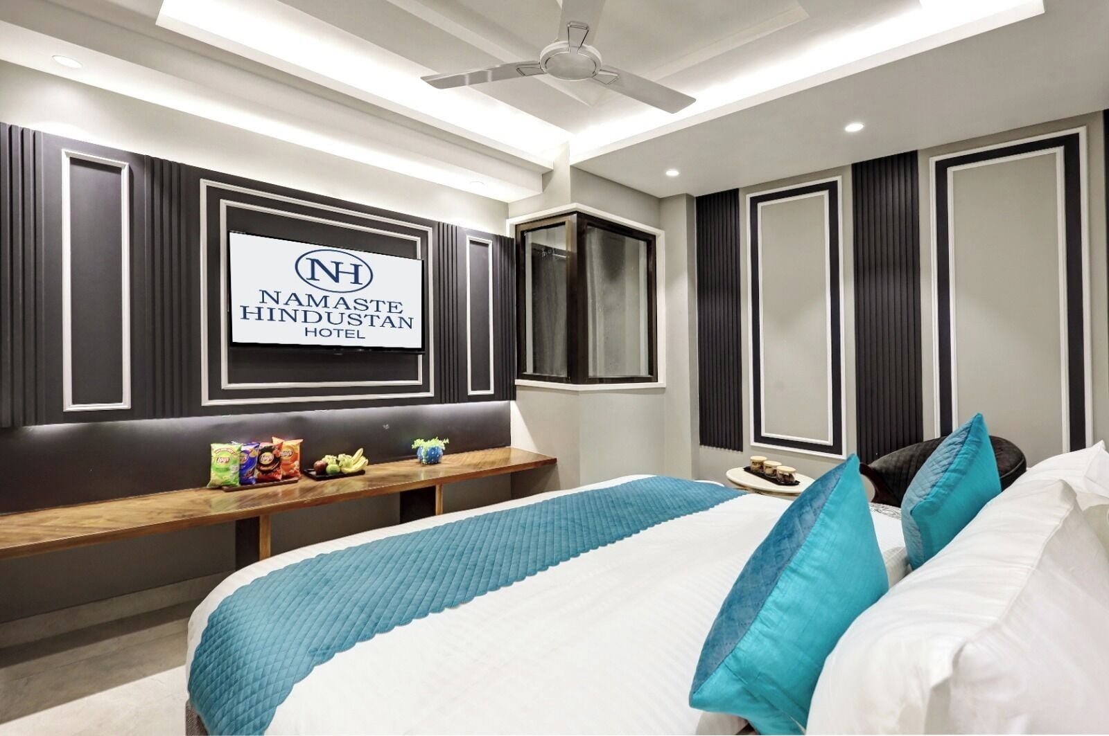 A hotel room with a bed, a bedside table with snacks, a TV mounted on the wall, and a ceiling fan.