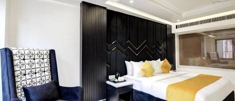 A modern hotel room with a large bed, a dark accent wall, a seating area with a chair, and a small side table.